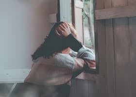 A woman sits indoors gazing thoughtfully out of a window, evoking emotions of loneliness and reflection.